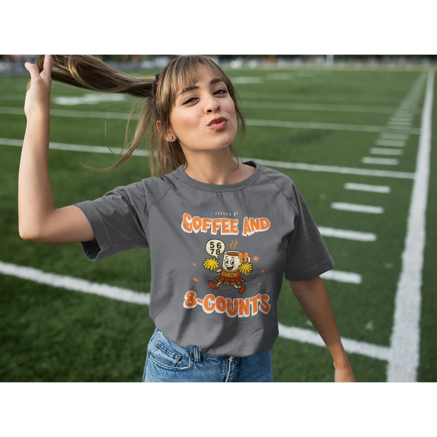 Cheerleading coach wearing a grey t-shirt with a cheerleading graphic design on a football field