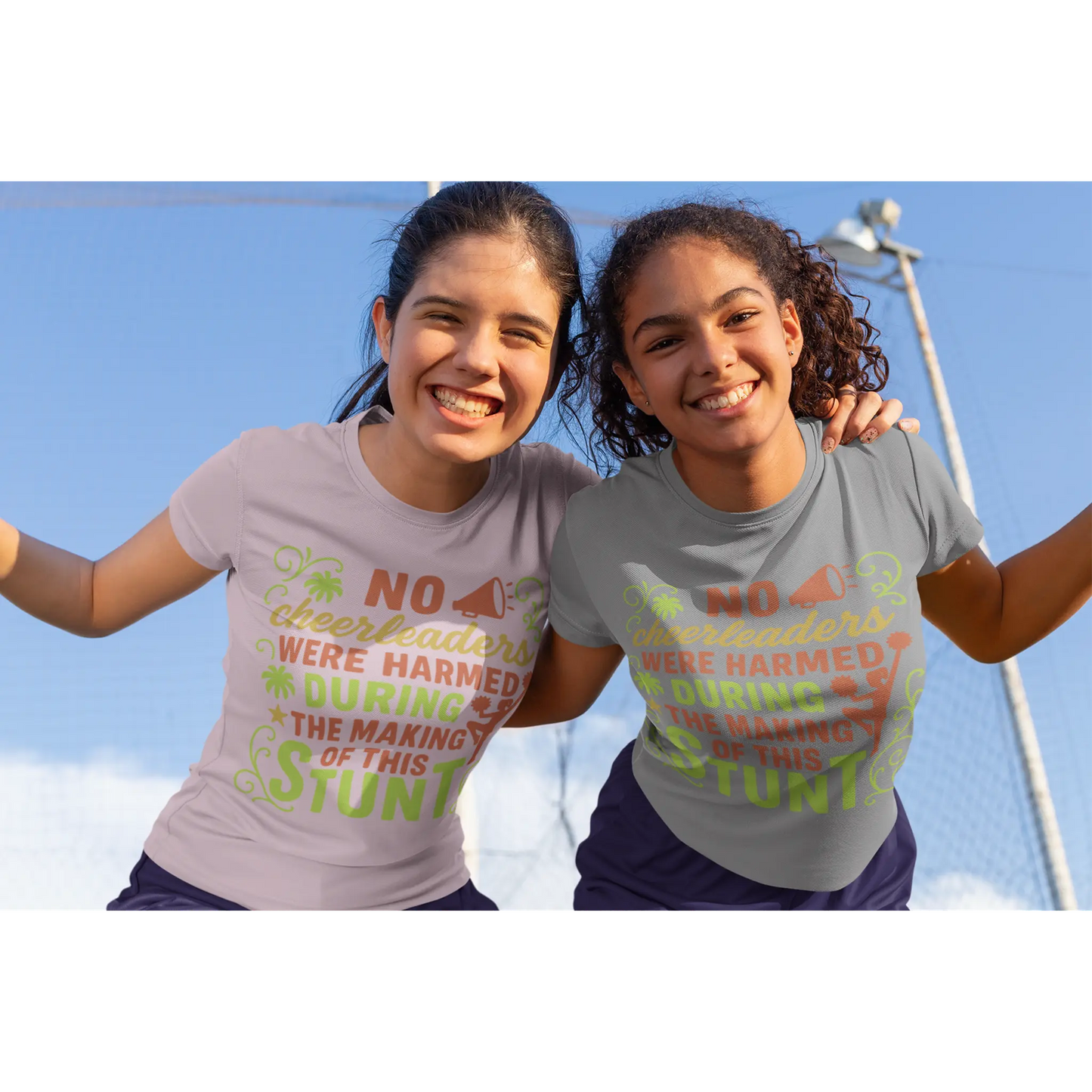 Two girls wearing t-shirts with humorous cheerleading text against a clear blue sky.