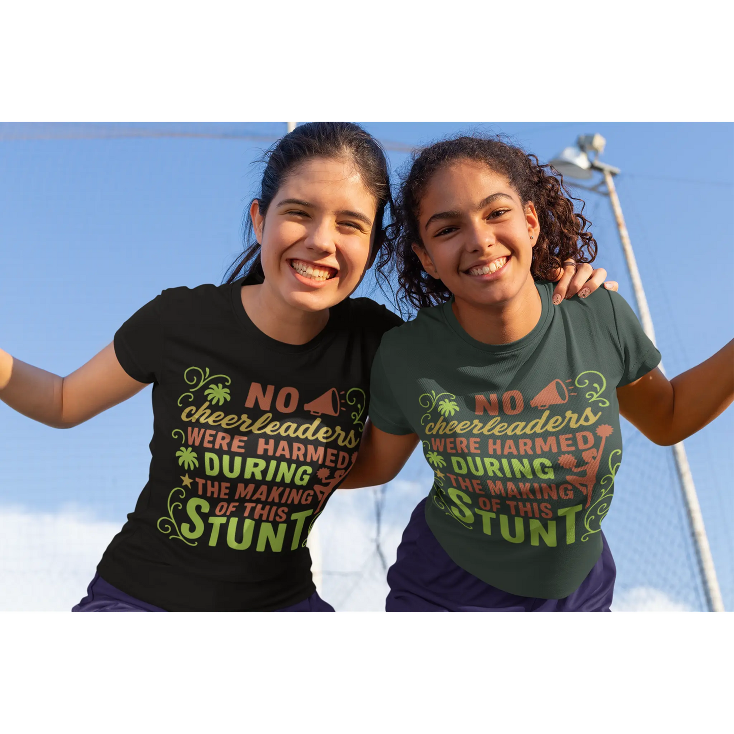 Two girls wearing t-shirts with a humorous message about cheerleaders, standing outdoors against a clear blue sky.