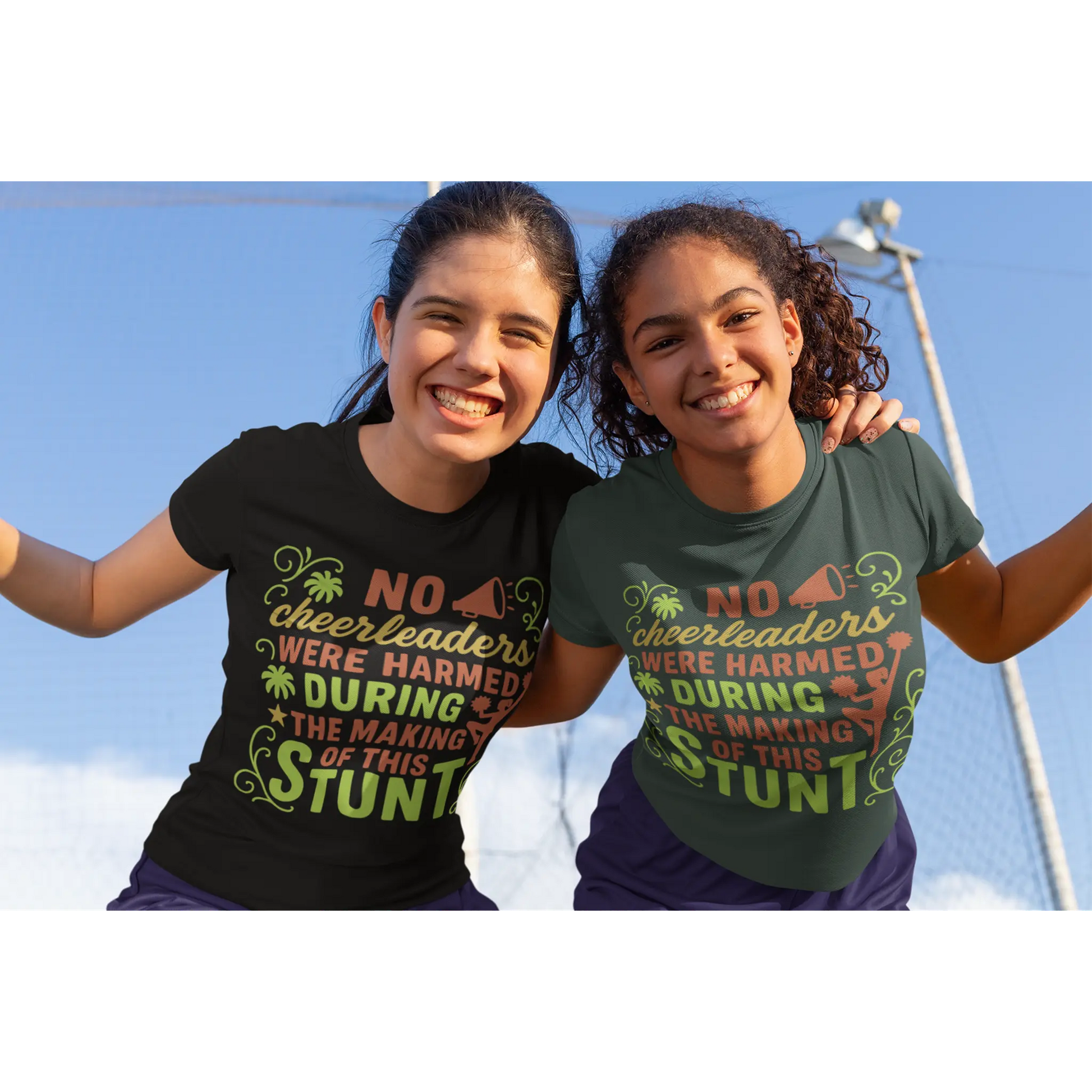 Two girls wearing t-shirts with a humorous message about cheerleaders, standing outdoors against a clear blue sky.